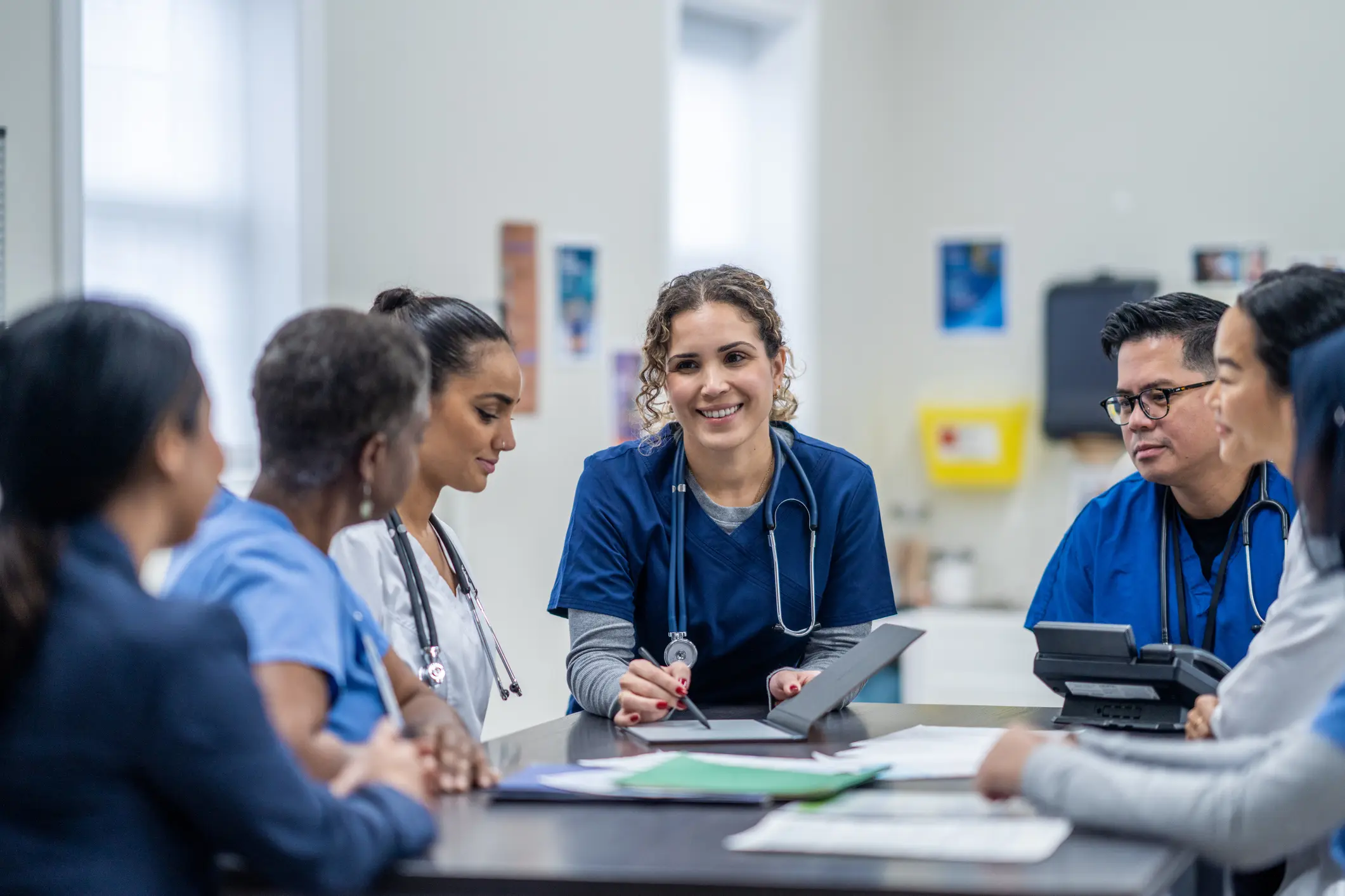 A small group of medical professionals gather around a table as they meet together to discuss patient cases.  They are each dressed professionally and have files scattered between them for reference.
